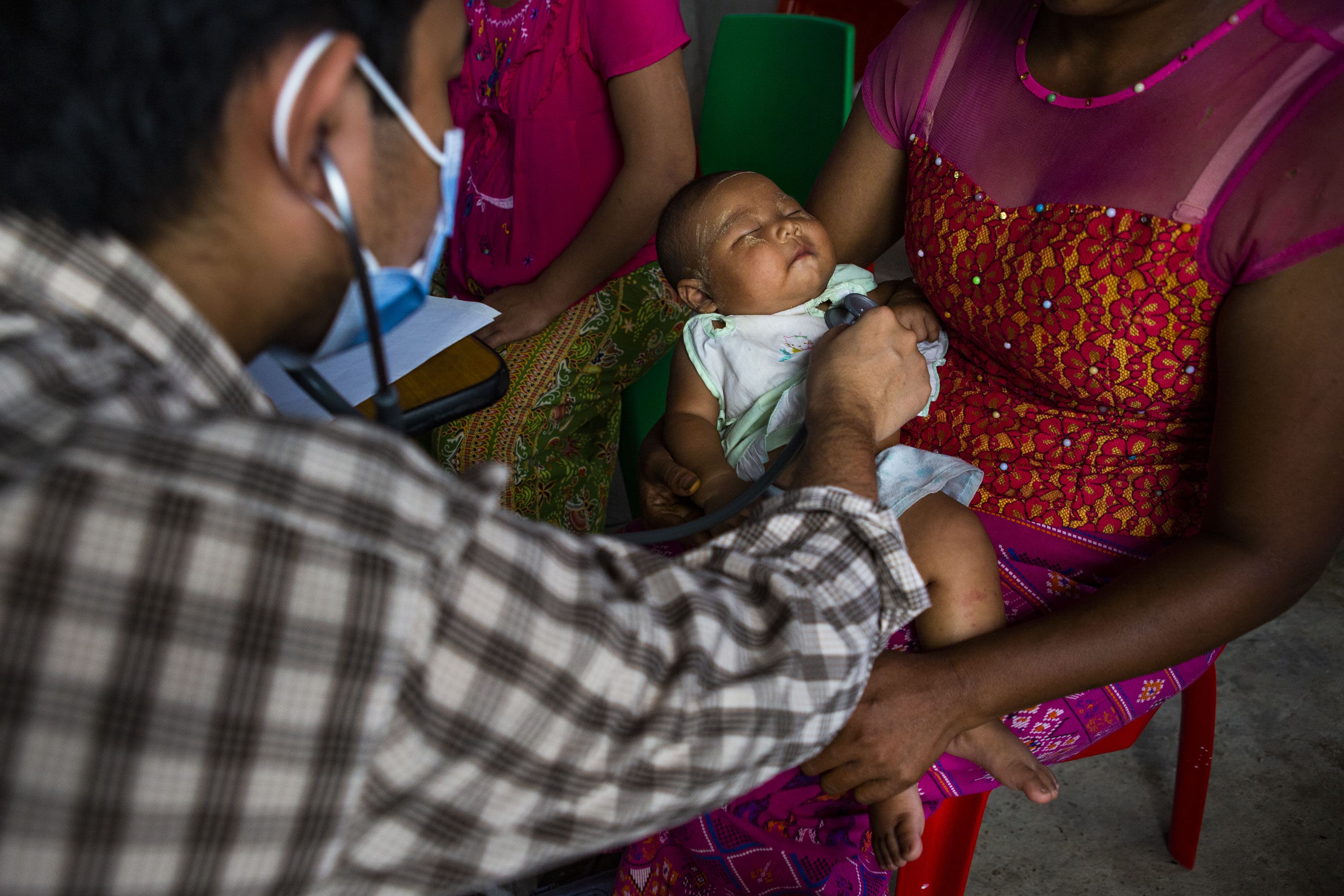 A baby is examined by a Tdh doctor in Myanmar. A doctor, photographed from behind, stretches out his arm to examine a sleeping baby lying on his mother's legs.