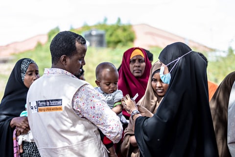 A Tdh staff member carries a baby who is having their arm measured by another person.