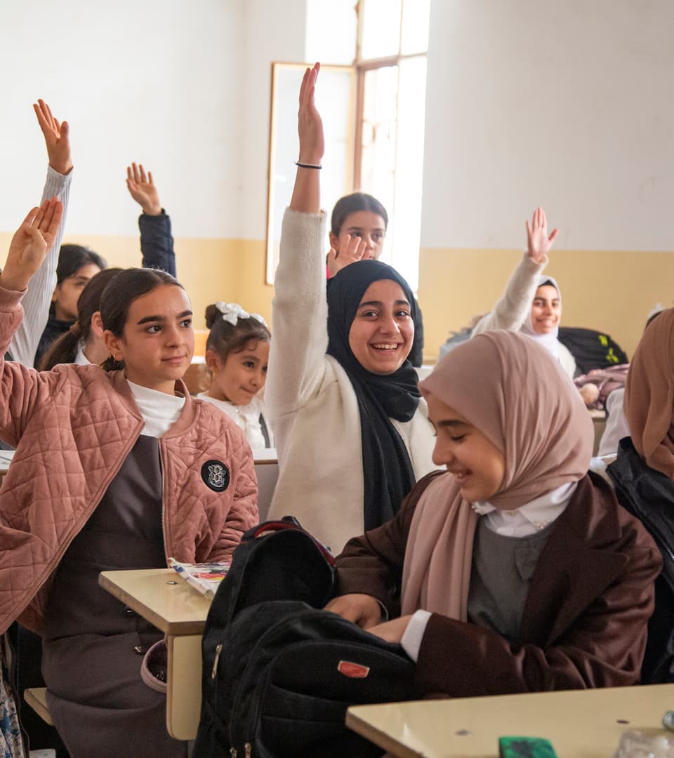 In a classroom, several girls sit at their desks and raise their hands.