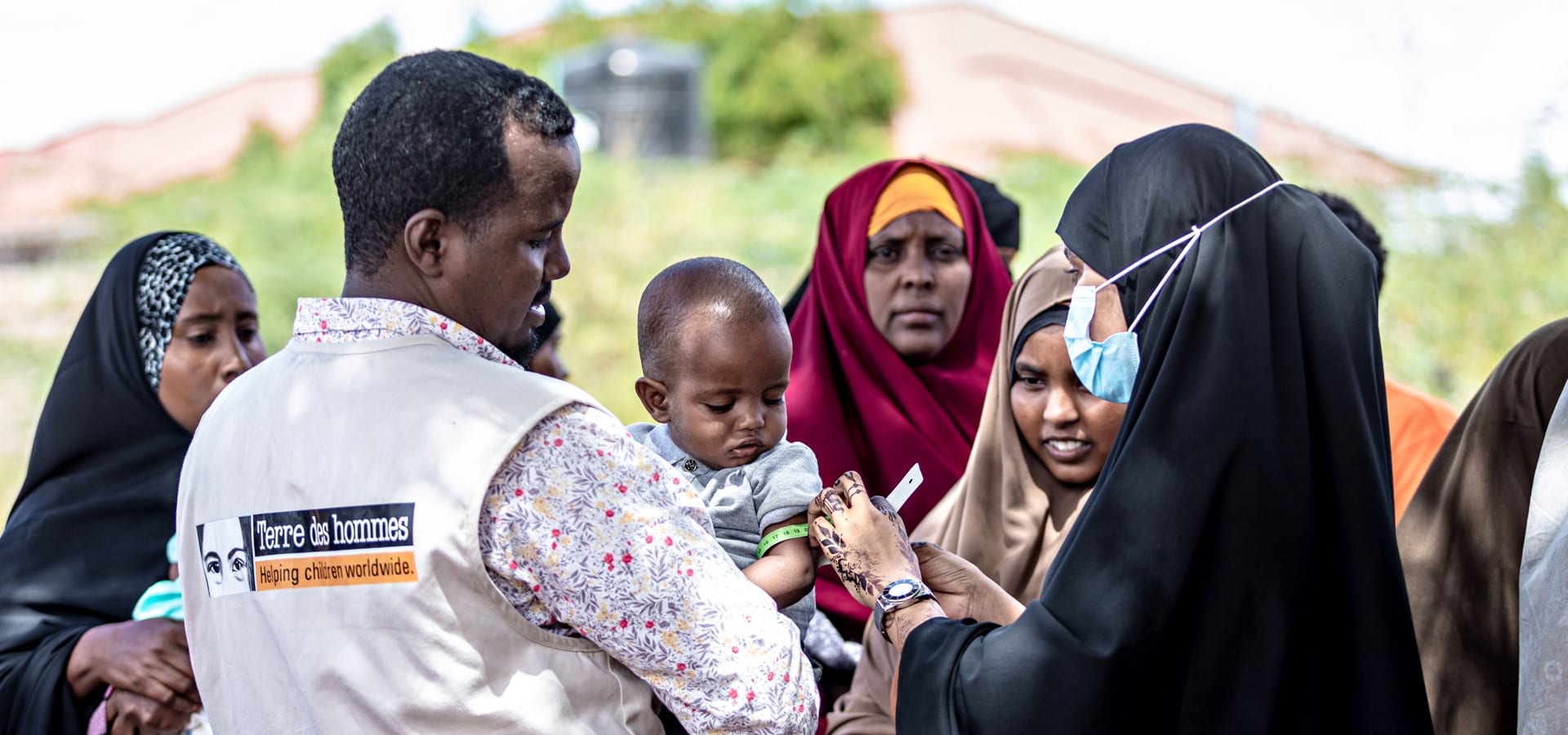 A Tdh staff member carries a baby who is having their arm measured by another person.