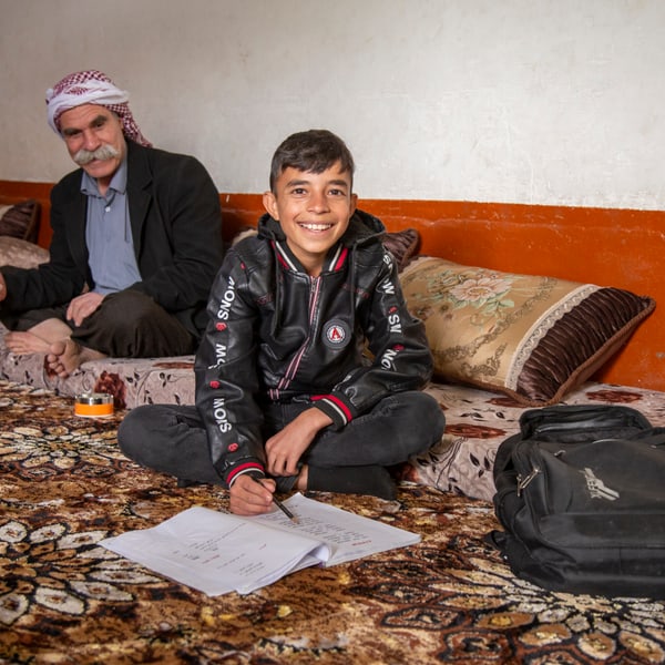 Young Zidan, sitting on the ground, smiles at the camera. His father is sitting behind him.