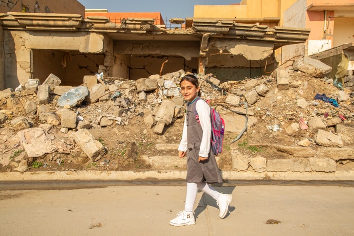 A girl carrying a backpack walks to the left past a ruined building.