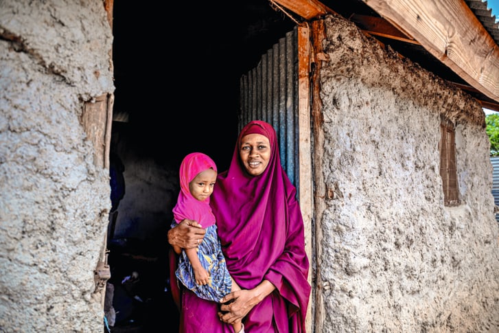 Sahara, die in Purpur verschleiert ist, trägt ihre Tochter Maryan auf dem Arm vor der Tür ihres Hauses.