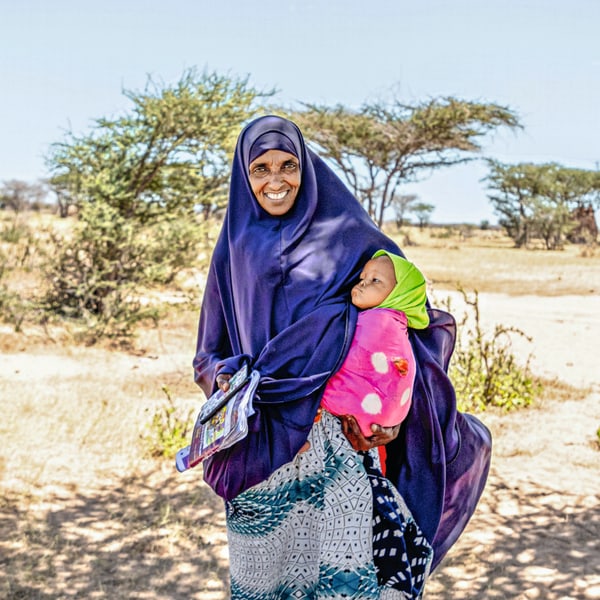 Kamila, veiled in purple, stands in the middle of a dry field, holding her niece draped in pink on her hip.