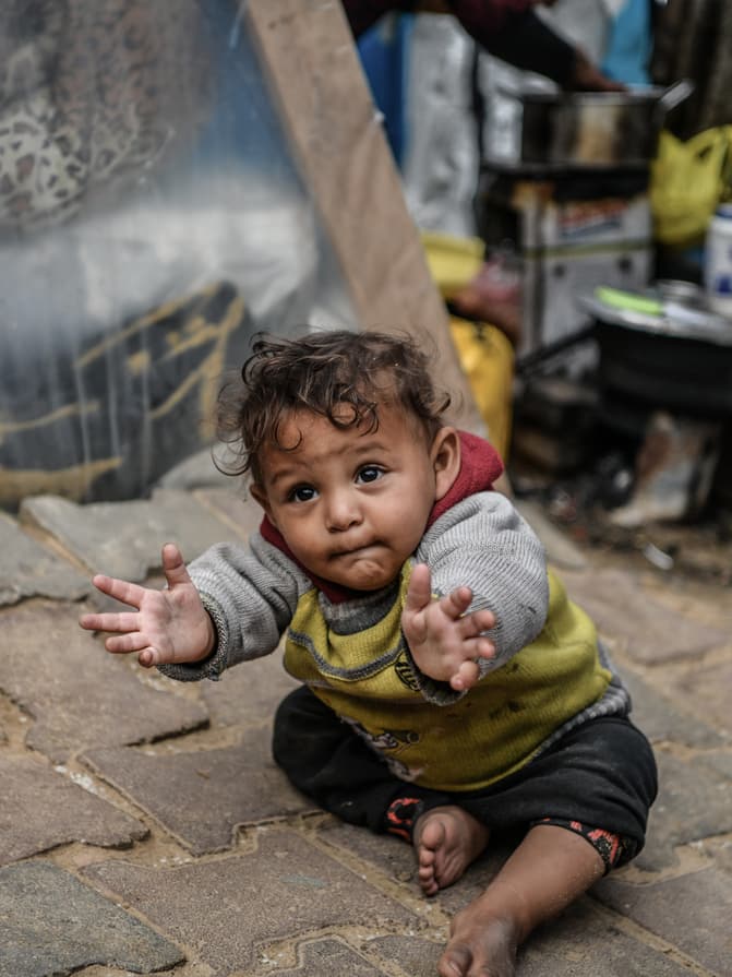 A baby sits on the ground in front of a tent, their arms outstretched towards the camera.